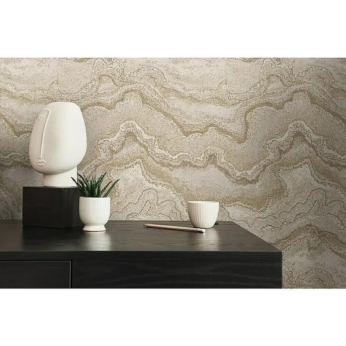 A.S. Création Stories of Life Vliestapete Marmoroptik Metallic-Effekt - Sandcreme, Steinoptik, 10,05 x 0,53 m - Dekoratives Stillleben mit einer weißen Skulptur, einer Pflanze, einer Tasse und einem Bleistift auf einer dunklen Kommode vor einer gemusterten Wand.