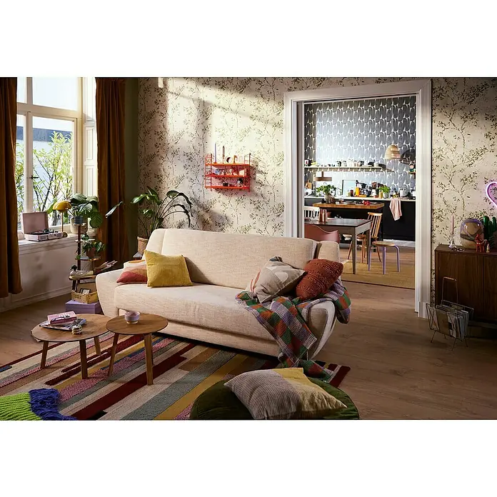 A.S. Création Stories of Life Vliestapete Zweig - Beige-Grün-Gold, Floral, 10,05 x 0,53 m - Gemütliches Wohnzimmer mit einem hellen Sofa, bunten Kissen und einem farbenfrohen Teppich, das zum Entspannen einlädt. Die Einrichtung ist stilvoll und einladend.