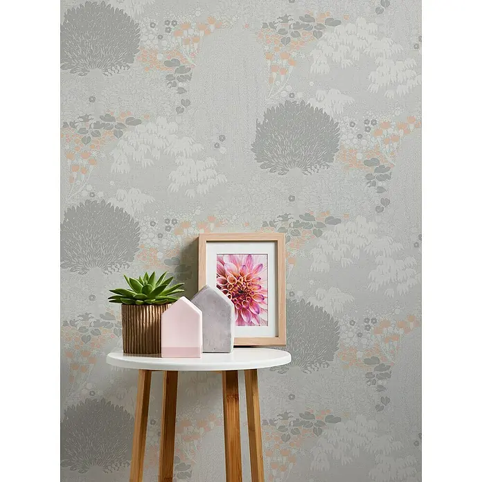 A.S. Création Famous Garden Vliestapete - Hellgrau-Weiß-Rosa, Floral, 10,05 x 0,53 m - Nahaufnahme eines Tisches mit Topfpflanze, Bilderrahmen und dekorativen Häusern vor einer floralen Tapete.