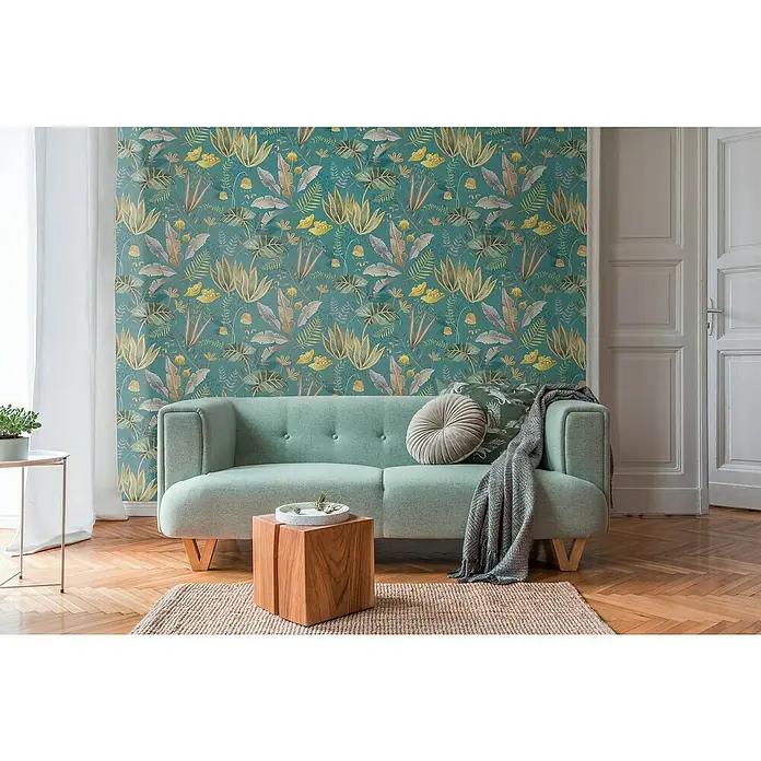 A.S. Création Famous Garden Vliestapete - Petrol-Grün-Gelb, Floral, 10,05 x 0,53 m - Ein mintgrünes Sofa mit einem runden Kissen und einer Decke vor einer grünen, floral gemusterten Wand.
