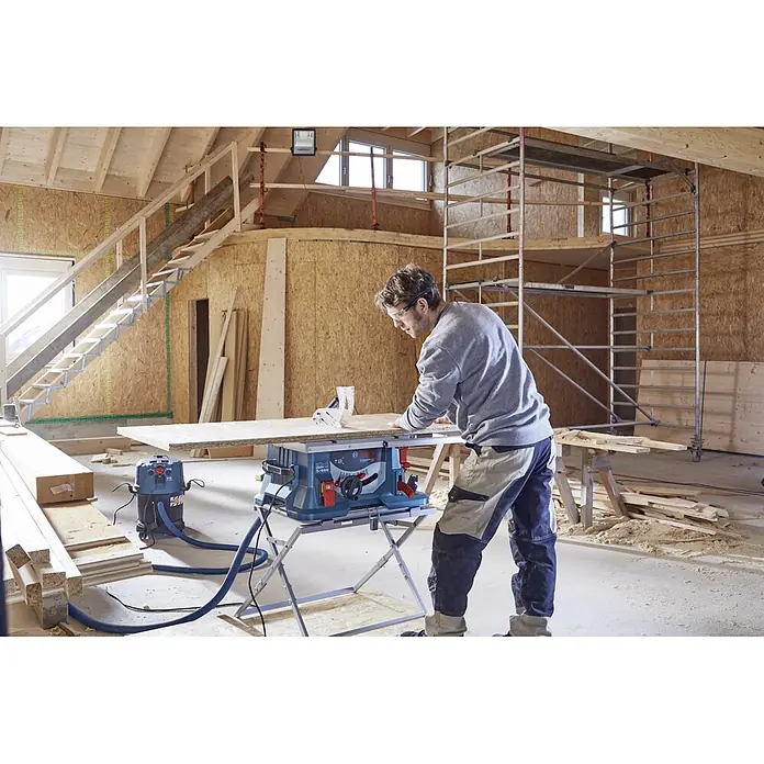 Bosch Professional Tischkreissäge GTS 635-216 - 1.600 W, Durchmesser Sägeblatt: 216 mm, Schnitttiefe bei 90 °: 70 mm, Ohne Untergestell - Ein Mann, der einen Tisch mit einer Bosch Kreissäge bearbeitet, in einer Werkstatt mit viel Holz und einer Treppe im Hintergrund.