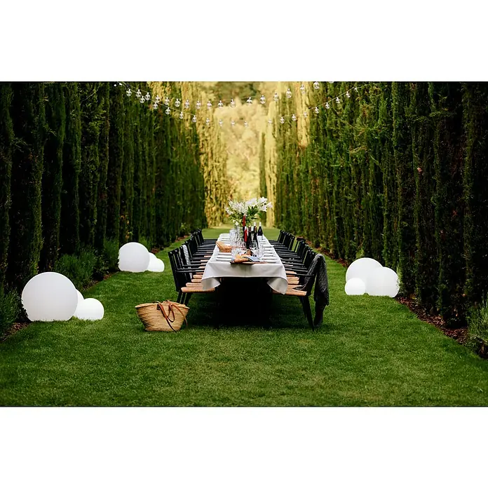 New Garden Design-Außenleuchte Buly 20 - Durchmesser: 20 cm, E27, IP65 - Im Freien ist ein langer Esstisch mit Stühlen für ein festliches Abendessen in der Natur vorbereitet, umgeben von Lichtern und Bäumen.