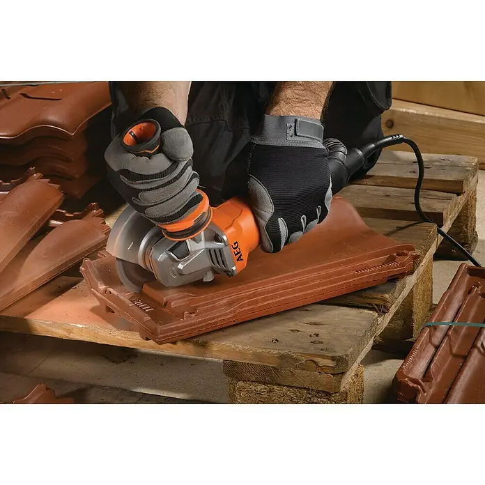 AEG Powertools Winkelschleifer-Set WS 13-125 SXE - 1.300 W, 125 mm - Nahaufnahme des AEG Winkelschleifers beim Schneiden einer Dachziegel, getragen durch Schutzhandschuhe, auf einer Holzpalette.