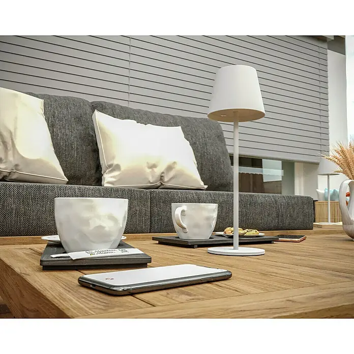 Starlux Paola Akku-Tischleuchte - 3 W, L x B x H: 11 x 11 x 36 cm, Weiß, Warmweiß - Diese Nahaufnahme zeigt eine elegante Tischlampe mit zwei Kaffeetassen und Smartphones auf einem Holztisch vor einer grauen Couch und Jalousien.