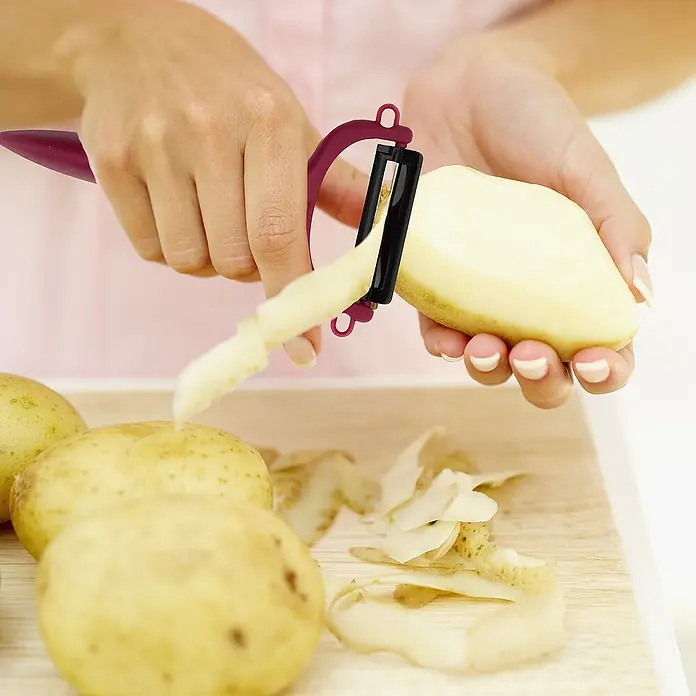 Pelador de verduras - Hoja de cerámica, Beige/Blanco/Negro/Gris/Burdeos, 1 pzs. - Un pelador de verduras en acción, pelando una patata, con más patatas sobre una tabla de cortar al fondo.