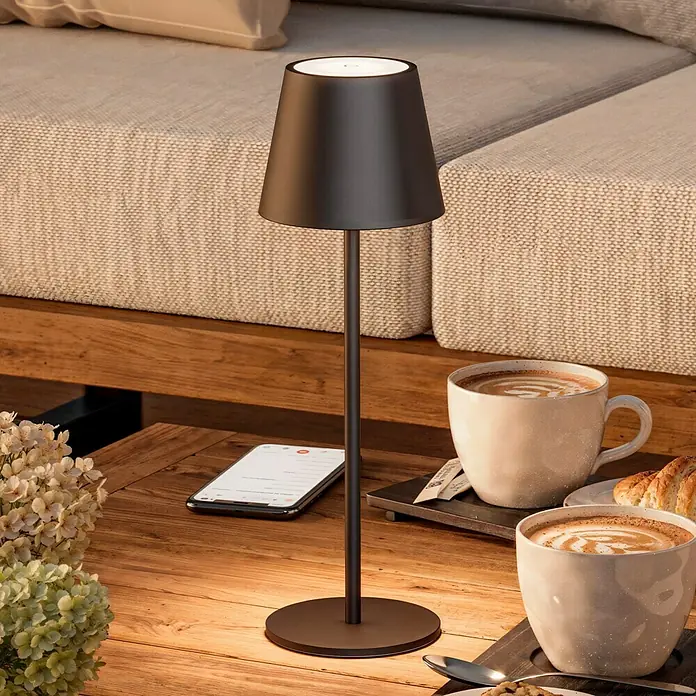 Starlux Paola Akku-Tischleuchte - 3 W, L x B x H: 11 x 11 x 36 cm, Schwarz, Warmweiß - Elegante schwarze Tischleuchte auf einem Holztisch mit Kaffeetassen und einem Smartphone. Stilvolle Beleuchtung für gemütliche Momente.