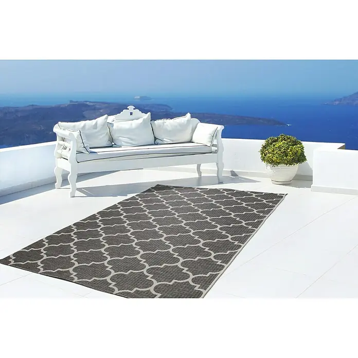 Outdoor-Teppich Sunset - Grau, 170 x 120 cm, 100 % Polypropylen - Ein grauer Teppich mit marokkanischem Muster liegt auf einer Terrasse mit Meerblick und Sitzmöbeln.