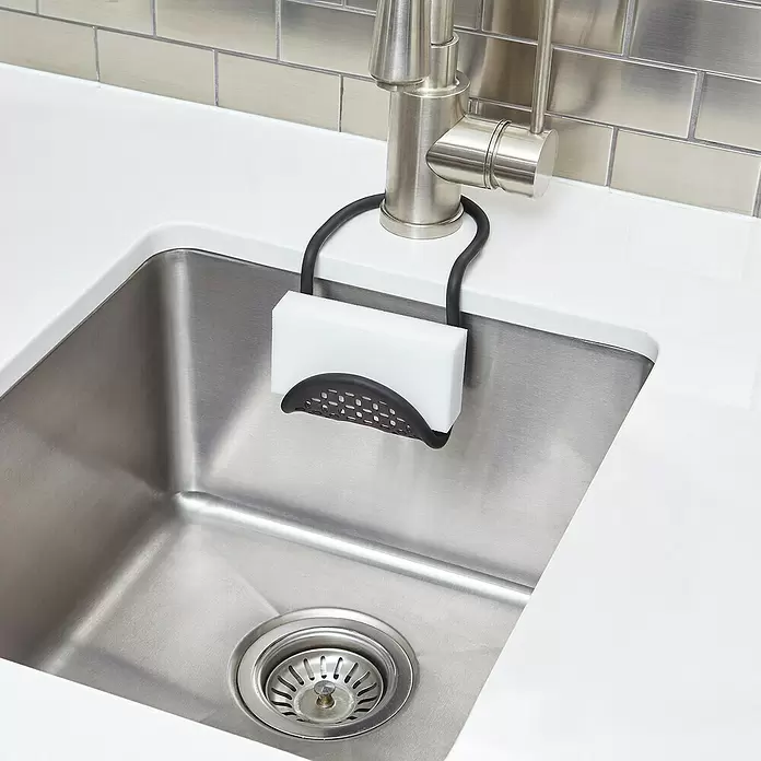 Umbra Spülbecken-Organizer Sling - 26 cm x 11 mm x 1 cm, Schwarz - Schwarzer Schwammhalter mit weißem Schwamm, hängt am Wasserhahn in einer Edelstahlspüle.