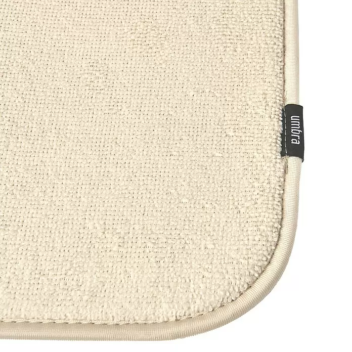 Umbra Abtropfmatte Udry - L x B x H: 61 x 46 x 4 mm, Kunststoff, Beige - Ecke einer cremefarbenen, texturierten Trockenmatte mit schwarzem "umbra" Markenetikett.