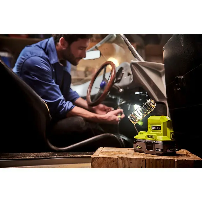 Ryobi ONE+ Soldador de puntos de batería R18SOI-0 - Apto para: Soldaduras finas - Lámpara de trabajo inalámbrica Ryobi ONE+ sobre tabla de madera mientras un hombre trabaja en el coche. La lámpara es amarilla y negra con batería de 3.0 Ah.