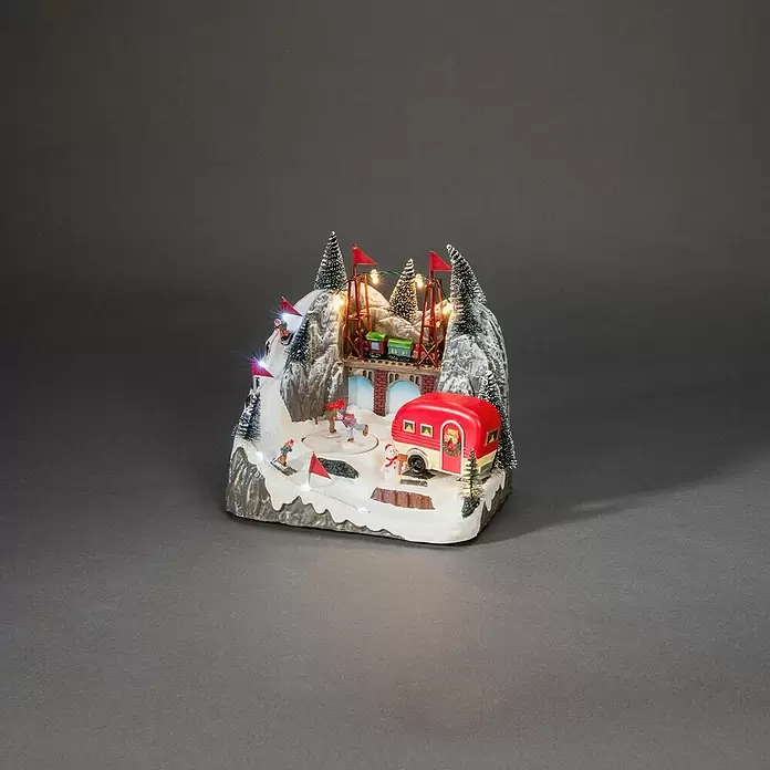 Konstsmide LED-Szenerie Caravan - L x B x H: 16 x 22 x 21,5 cm, Bunt, Batteriebetrieben, LED - Beleuchtete Weihnachtsdorf-Szene mit Skifahrern, einem Eisläufer, einem Zug auf einer Brücke und einem roten Wohnwagen in einer Schneelandschaft.