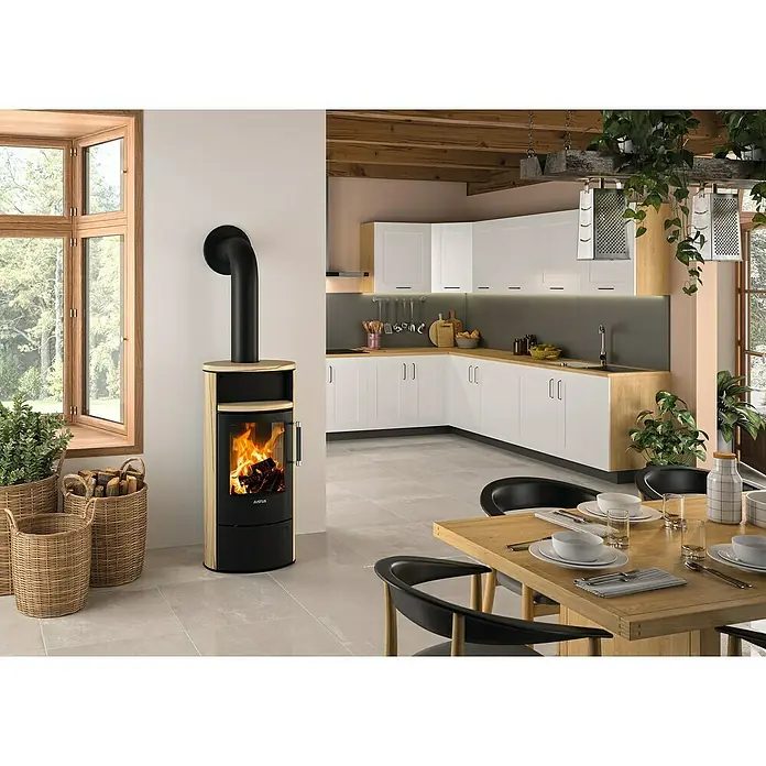 Justus Kaminofen Island 5 2.0 - 5 kW, Raumheizvermögen: 88 m³, Material Verkleidung: Speckstein, Sandstein/Schwarz - Gemütliches Esszimmer mit einem Justus Kaminofen, der Wärme und Stil verleiht. Helle Küche im Hintergrund.