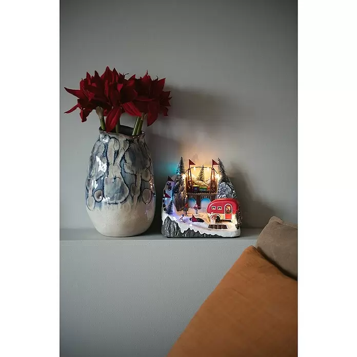 Konstsmide LED-Szenerie Caravan - L x B x H: 16 x 22 x 21,5 cm, Bunt, Batteriebetrieben, LED - Eine Vase mit roten Blumen neben einer beleuchteten Weihnachtsdorf-Szene mit Skifahrern und einem roten Wohnwagen auf einem Regal.