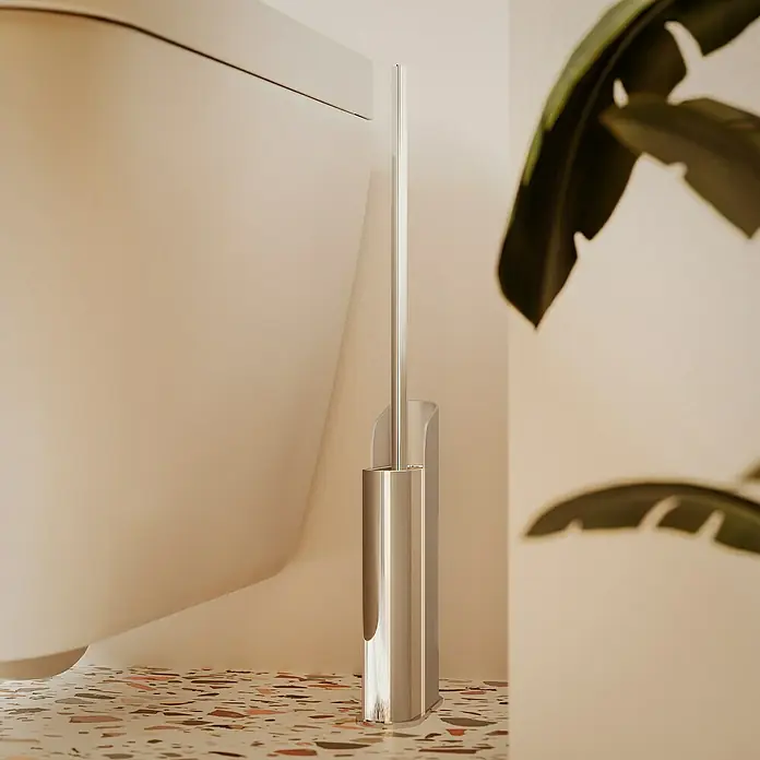 Cosmic Escobillero de suelo y pared Slim - Silicona, Cromo - Un moderno portaescobillas de baño plateado se encuentra junto a un inodoro sobre un suelo de terrazo. El diseño es elegante y minimalista.