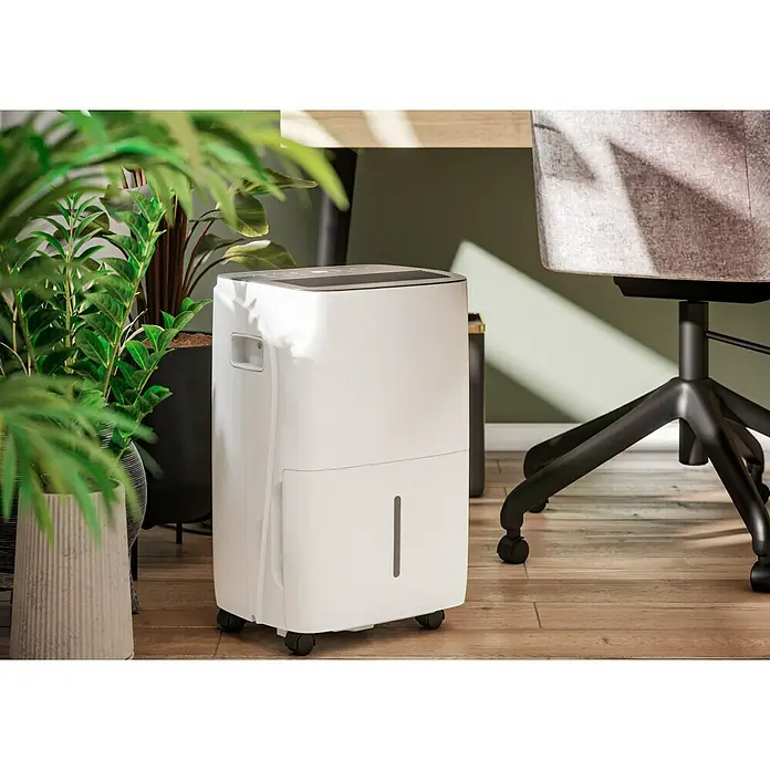 Voltomat Luftentfeuchter - Entfeuchtungsleistung: 60 l/Tag, Weiß/Grau, 900 W, Raumvolumen: 250 m³ - Ein weißer Luftentfeuchter mit Rollen steht auf einem Holzboden neben Pflanzen und einem Bürostuhl. Praktisches Design für ein angenehmes Raumklima.