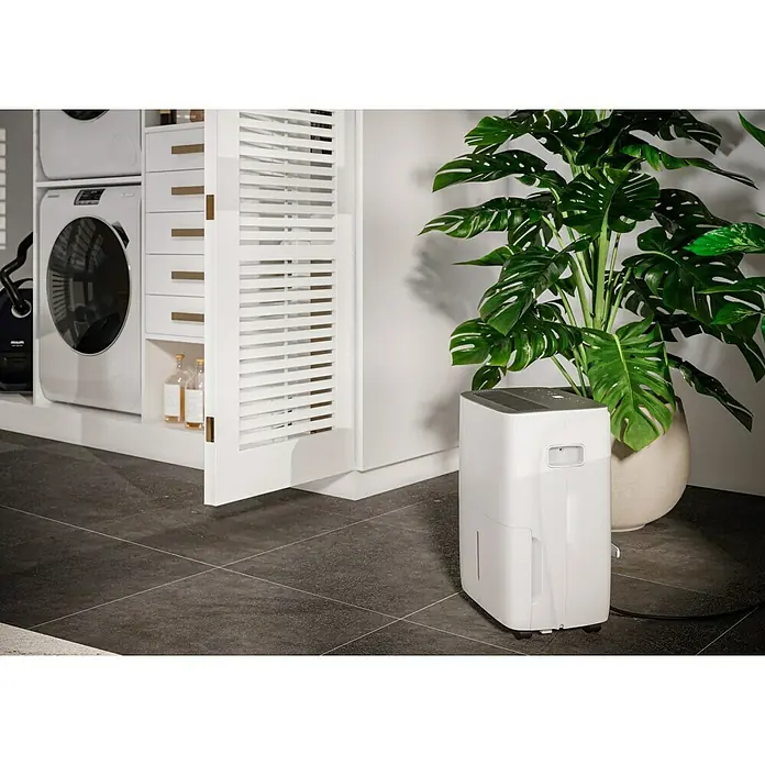 Voltomat Luftentfeuchter - Entfeuchtungsleistung: 40 l/Tag, Weiß/Grau, 680 W, Raumvolumen: 200 m³ - Ein weißer Luftentfeuchter steht neben einer großen Zimmerpflanze in einem Raum mit dunklen Fliesen. Er bietet eine effiziente Lösung für Feuchtigkeit.