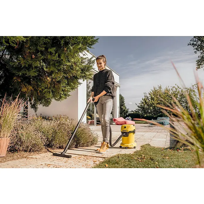 Kärcher Aspirador en seco y húmedo eléctrico WD 3 V-17/4/20 - 1.000 W, Volumen de recipiente: 17 l - Una mujer usa una aspiradora Kärcher amarilla para sólidos y líquidos en el exterior para quitar las hojas. La aspiradora se encuentra en una zona pavimentada al lado de verde.
