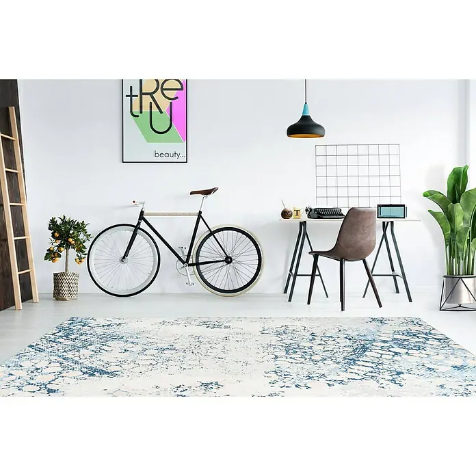 Kayoom Flachgewebeteppich Galaxy - Creme/Blau, 150 x 80 cm, 100 % Polyester - Ein strukturierter Teppich mit blau-weißen Akzenten liegt in einem stilvollen Raum mit Fahrrad und Schreibtisch.