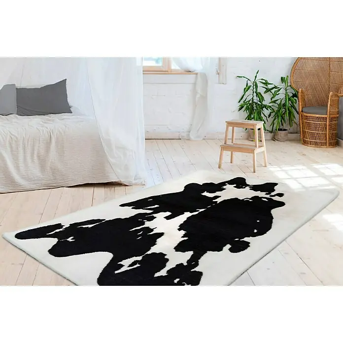 Kayoom Hochflorteppich Kuh - Weiß/Schwarz, 160 x 120 cm, 100 % Polyester - Ein weißer Teppich mit schwarzen Flecken im Kuhfellmuster liegt in einem hellen Raum mit Holzboden und einem Bett im Hintergrund.