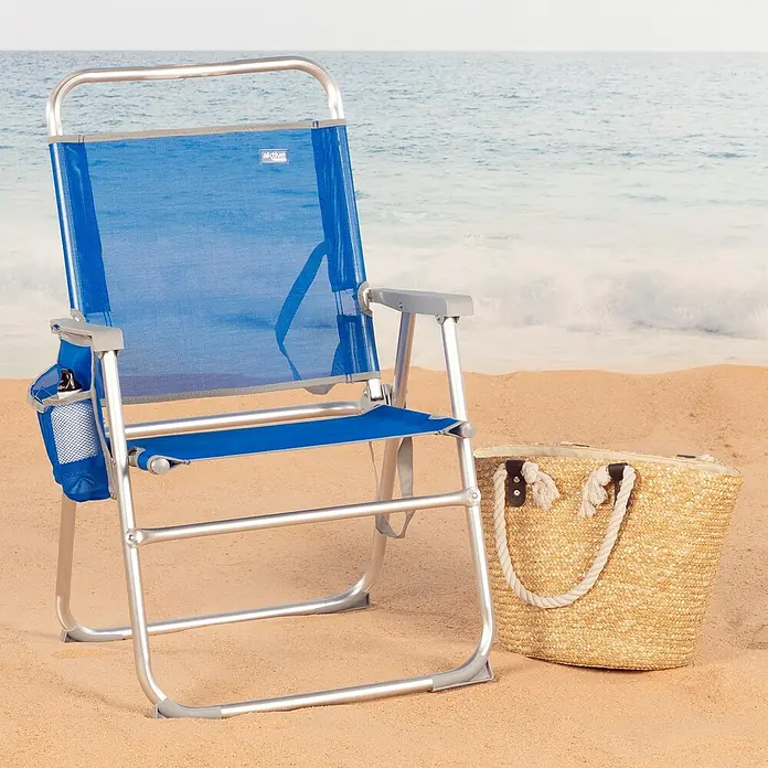 Silla de camping Gomera - 56 x 50 x 88 cm, Azul, Aluminio/Textiline - Una silla de playa azul de aluminio con portavasos y un bolso de playa de paja están sentados en la playa.
