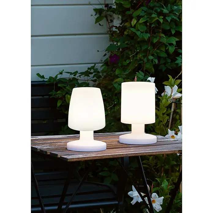 Reality Leuchten Akku-Tischleuchte Dora - 1,5 W, L x B x H: 13 x 13 x 21 cm, Weiß, Warmweiß - Zwei weiße, leuchtende Tischlampen stehen auf einem Holztisch im Freien, umgeben von üppigem Grün, und sorgen für eine gemütliche Atmosphäre.