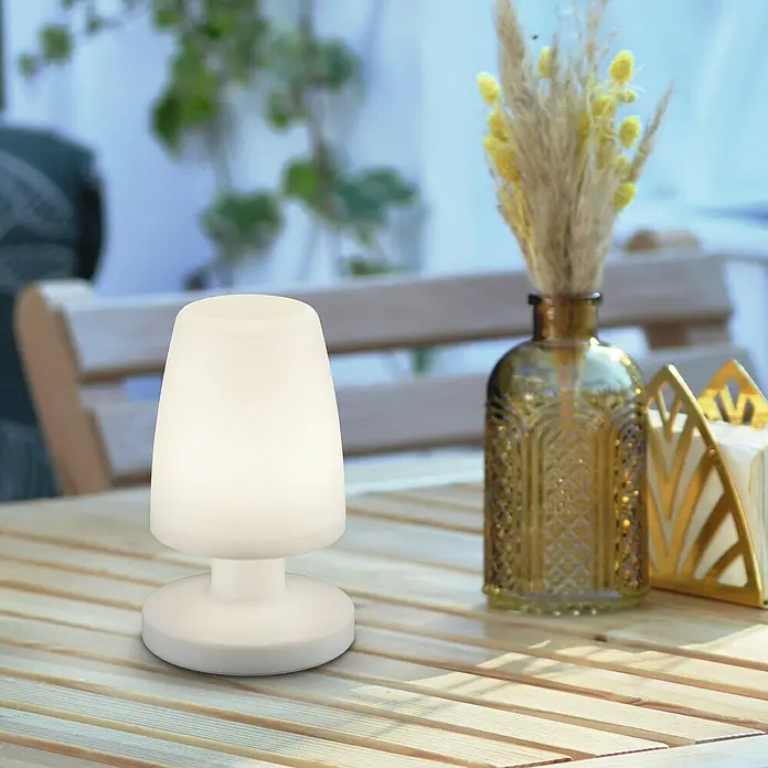 Reality Leuchten Akku-Tischleuchte Dora - 1,5 W, L x B x H: 13 x 13 x 21 cm, Weiß, Warmweiß - Stimmungsvolle Tischleuchte mit warmweißem Licht, perfekt für den Außenbereich, neben einer Blumenvase auf einem Holztisch.