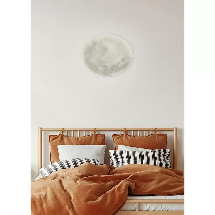 Trio Lighting Plafón LED redondo Luna - 22 W, Ø x Al: 40 x 10 cm, Gris - Una lámpara de luna blanca realista cuelga sobre una cama con cojines de terracota y rayas.