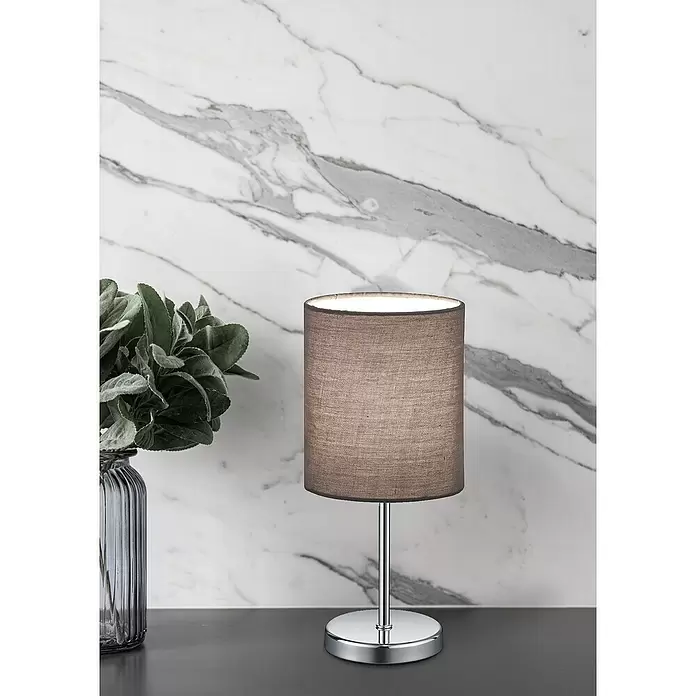 Reality Leuchten Tischleuchte rund Jerry - 40 W, Ø x H: 130 mm x 28,5 cm, Chrom, E14 - Tischlampe mit dunkelbraunem Stoffschirm und Chromfuß, auf einem Tisch neben einer Pflanze vor einer Marmorwand.