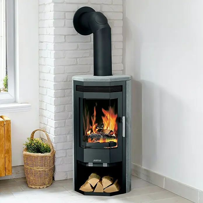 Justus Kaminofen Usedom 5 II - 5,5 kW, Raumheizvermögen: 88 m³, Material Verkleidung: Speckstein, Schwarz/Speckstein - Nahaufnahme eines Justus Kaminofens mit brennendem Holz und Holzscheiten im unteren Fach, neben einem Korb mit Pflanzen.