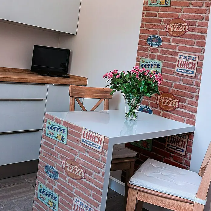 A.S. Création 3D-Designpanel pop.up - Beige/Braun/Creme, 52 cm x 2,5 m, Coffee - Esstisch mit dekorativer Backsteinoptik und Blumen in einer Vase. Die Stühle sind aus hellem Holz.