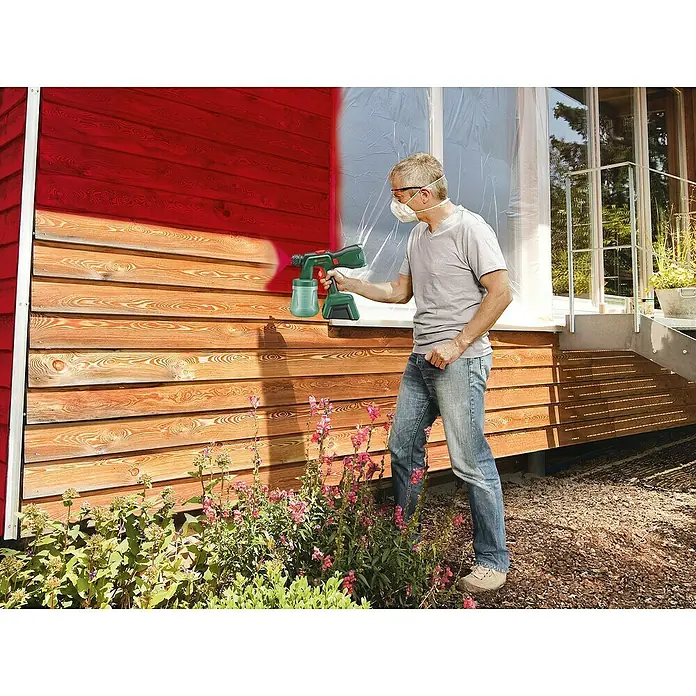 Bosch Power for All 18V Akku-Farbsprühsystem Universal Spray 18V-300 - 18 V, Ohne Akku, Max. Fördermenge: 0,3 l/min - Ein Mann streicht mit einer Farbspritzpistole ein Holzhaus rot. Die Fenster sind abgedeckt. Er trägt eine Maske.