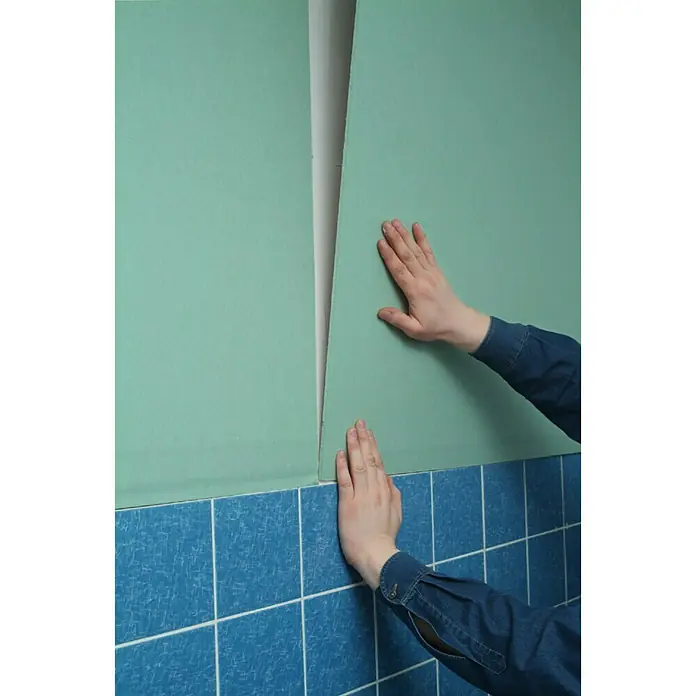 Knauf Gipskartonplatte GKBI - 1.250 x 900 x 6,5 mm, Imprägniert - Eine Nahaufnahme von Händen, die grüne Gipskartonplatten an einer Wand mit blauen Fliesen anbringen, die ein Baumaterial darstellen.