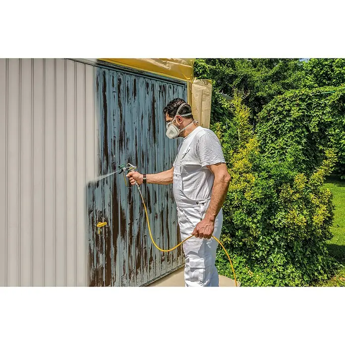 Wagner High Efficiency Airless Farbsprühsystem Power Painter 90 - 800 W, Förderleistung: 1,9 l/min - Mann lackiert mit einer Farbspritzpistole eine Garagentür, umgeben von Grün. Er trägt Schutzkleidung und eine Maske für die Arbeit.