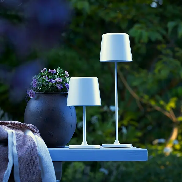 Brilliant Akku-Tischleuchte Kaami - 2 W, Weiß, Warmweiß - Zwei moderne Tischlampen in Weiß stehen auf einem Tisch im Freien, neben einem Topf mit lila Blumen und einer Decke.