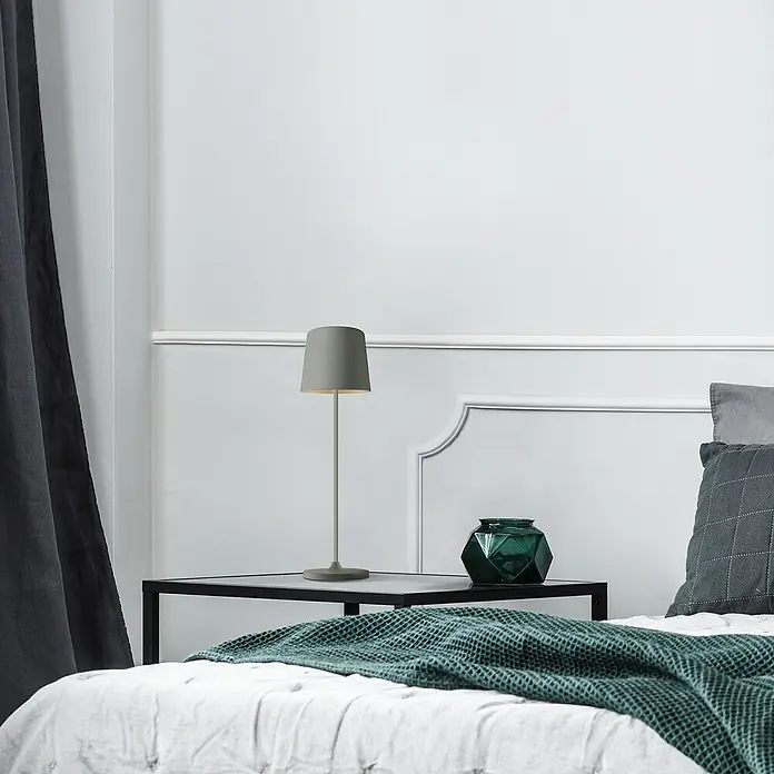 Brilliant Akku-Tischleuchte Kaami - 2 W, Grau, Warmweiß - Eine graue Lampe steht auf einem Nachttisch neben einem Bett mit grüner Decke und Kissen. Sie beleuchtet den Raum sanft.
