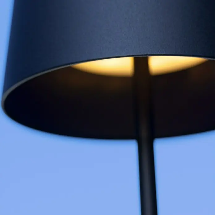 Brilliant Akku-Tischleuchte Kaami - 2 W, Schwarz, Warmweiß - Nahaufnahme einer stilvollen, modernen Lampe mit warmem Lichtschein vor blauem Hintergrund.