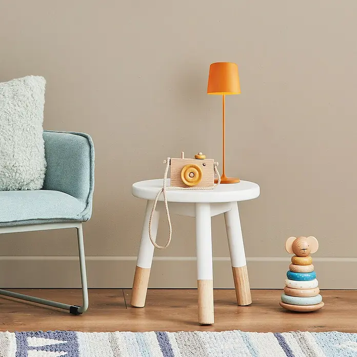 Brilliant Akku-Tischleuchte Kaami - 2 W, Orange, Warmweiß - Weißer Kinderhocker mit Holzbeinen, darauf eine Holzkamera und eine orangefarbene Lampe. Ein Spielzeugturm steht daneben.