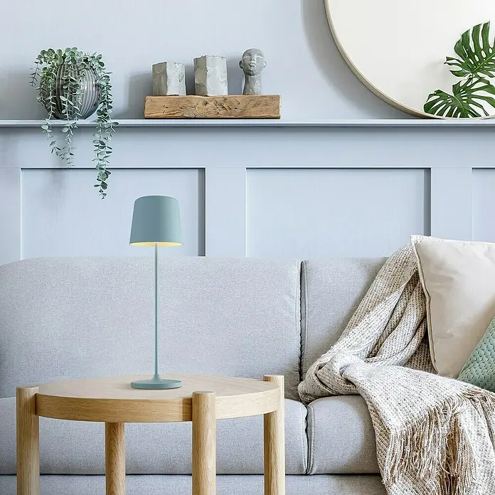 Brilliant Akku-Tischleuchte Kaami - 2 W, Mint, Warmweiß - Nahaufnahme einer hellblauen Tischleuchte auf einem runden Holztisch neben einem grauen Sofa mit Kissen und Überwurf.