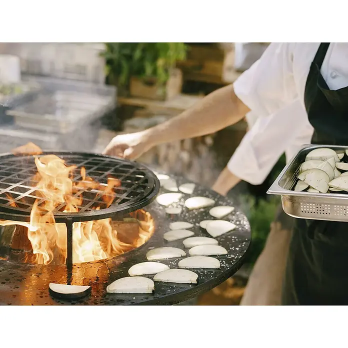Purline Barbacoa de carbón EFP56CO - Diámetro de la superficie de la parrilla: 98 cm, Óxido - Un chef asa rodajas de berenjena en una parrilla de fuego abierto, mientras sostiene más berenjena en un recipiente.