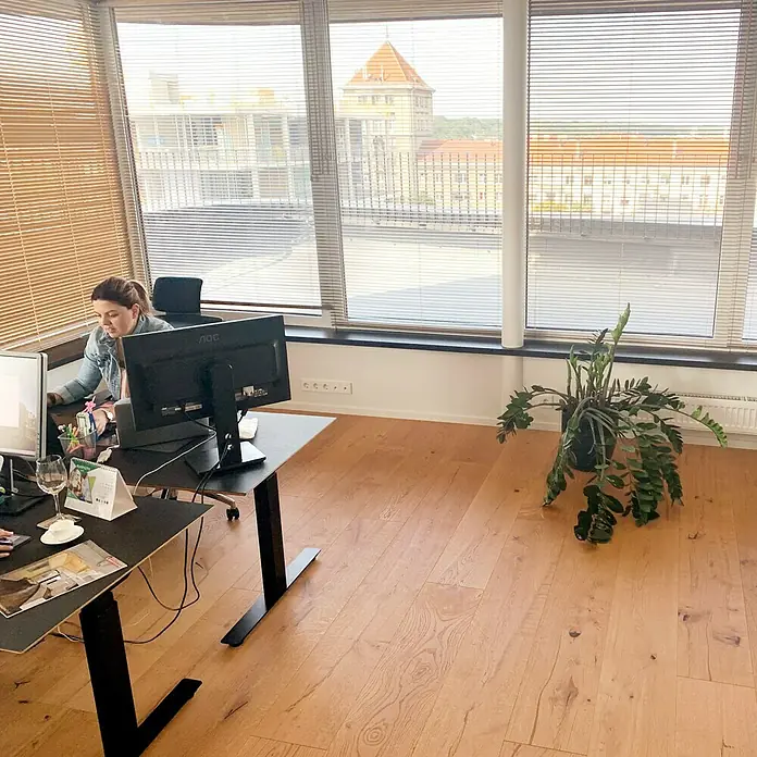 Parkettmuster Eiche Hotel - 280 x 160 x 15 mm, Landhausdiele - Eine Frau arbeitet an einem höhenverstellbaren Schreibtisch mit zwei Monitoren in einem hellen Büro mit Holzboden.