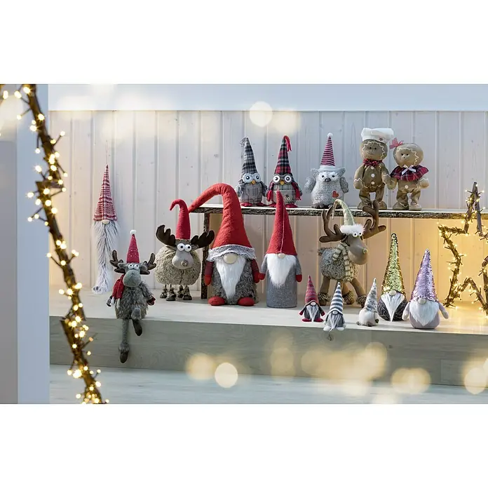 Weihnachtszwerg Weihnachtsmann mit langer Mütze - Textil, Weiß/Rot, Höhe: 52 cm - Festliche Szene mit Weihnachtszwergen, Elchen und Lebkuchenfiguren auf einem dekorierten Regal, umrahmt von Lichtern für eine warme, weihnachtliche Atmosphäre.