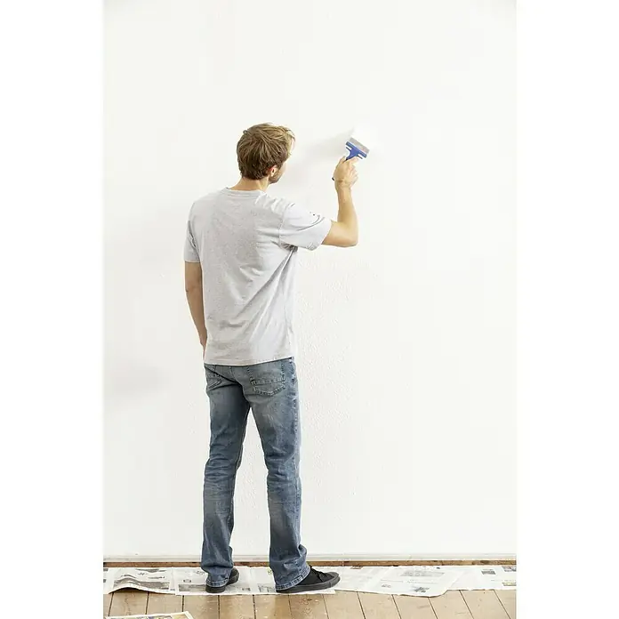 Knauf Easyputz-Spezialwerkzeug - Finishpinsel, Breite Borsten: 100 mm - Ein Mann streicht eine weiße Wand mit einem Pinsel, der blaue Akzente hat. Er trägt Jeans und ein graues T-Shirt und steht auf Zeitungspapier.