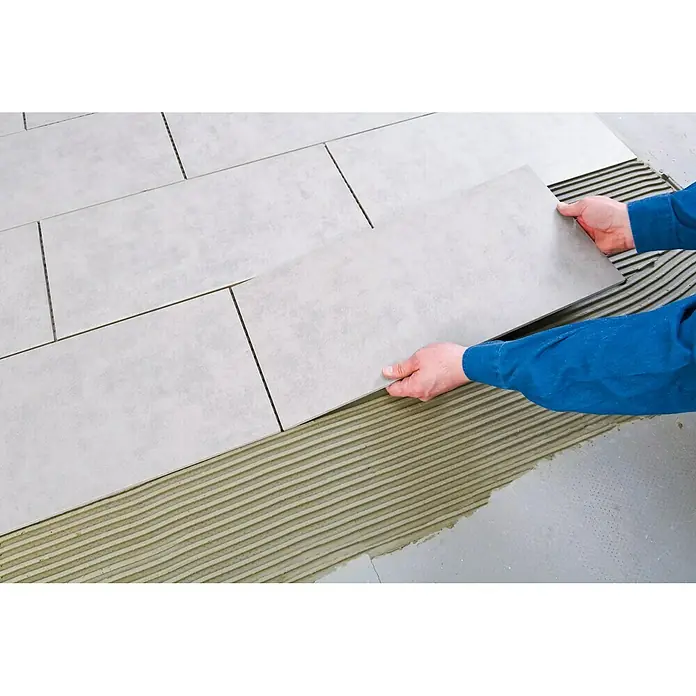 Knauf Flexkleber Extra - 5 kg - Das Bild zeigt einen Handwerker, der graue Bodenfliesen verlegt, wobei er sorgfältig eine Fliese in den vorbereiteten Kleber setzt.