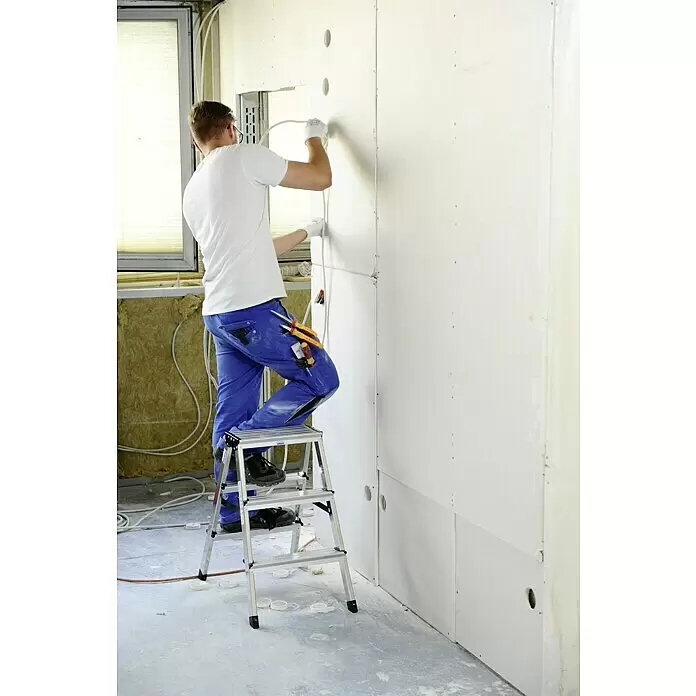 Krause Monto Escalera de tijera de doble acceso - Altura de trabajo: 2,2 m, Número de peldaños: 6 ud., Carga soportada: 150 kg, Aluminio - Trabajador sobre un taburete de dos peldaños plateado instalando cableado eléctrico en pared de pladur.