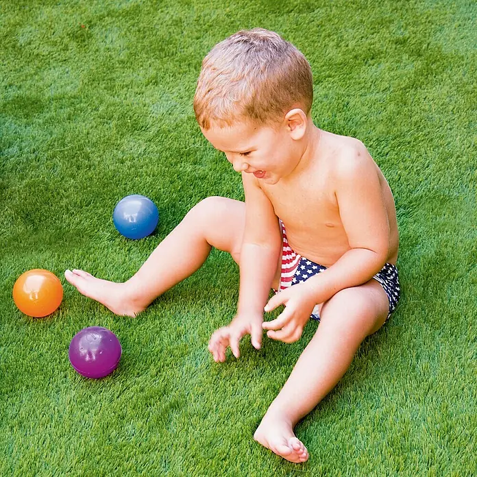 Césped artificial Paraíso - 5 x 1 m, Altura del pelo: 40 mm, 18.900 puntadas/m² - Un niño pequeño en traje de baño con bandera de EE. UU. juega con pelotas de colores en el césped y se divierte.