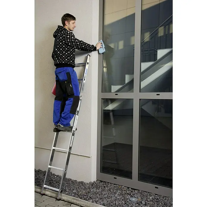 Krause Corda Escalera de apoyo - Número de peldaños: 8 ud., Altura de trabajo: 3,2 m, Carga soportada: 150 kg, Aluminio - Un hombre en una escalera limpia una ventana. Lleva ropa de trabajo azul y una chaqueta a cuadros. La escalera está sobre un lecho de grava frente al edificio.