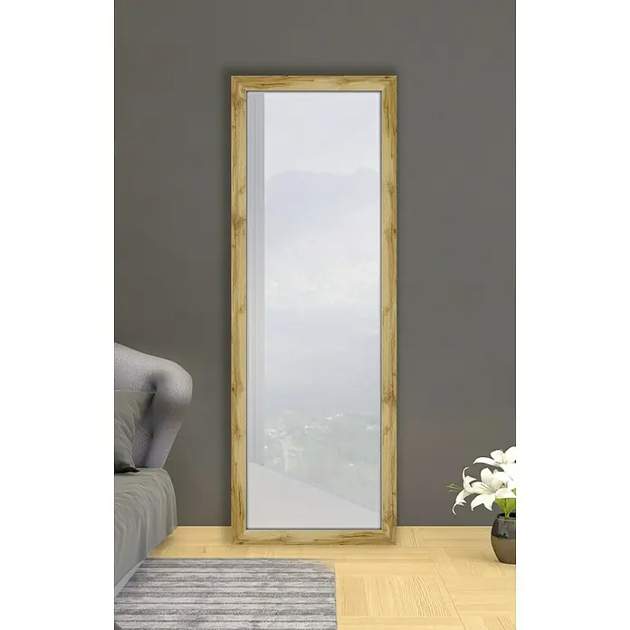 Espejo con marco de madera natural Mauren - 49 x 139 cm, Roble, Madera - Un espejo rectangular con marco dorado se apoya contra una pared gris, reflejando un paisaje borroso.