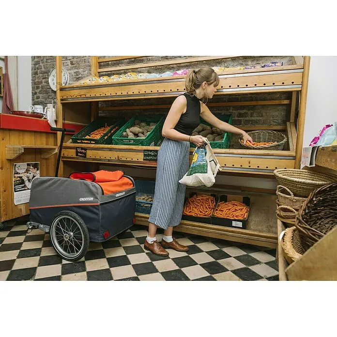 Croozer Fahrradanhänger Cargo Pakko - Traglast: 45 kg, Farbe: Campfire red - Eine Frau wählt Karotten aus einem Regal mit dem Croozer-Fahrradanhänger im Vordergrund.