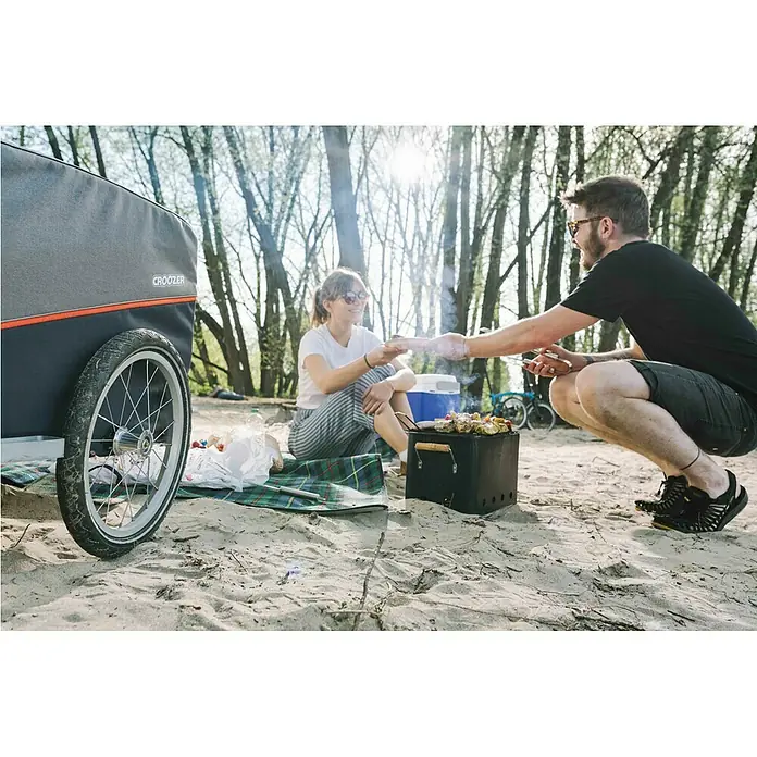 Croozer Fahrradanhänger Cargo Pakko - Traglast: 45 kg, Farbe: Campfire red - Ein Croozer Fahrradanhänger beim Picknick am Strand, wo ein Mann der Frau Grillgut anbietet.
