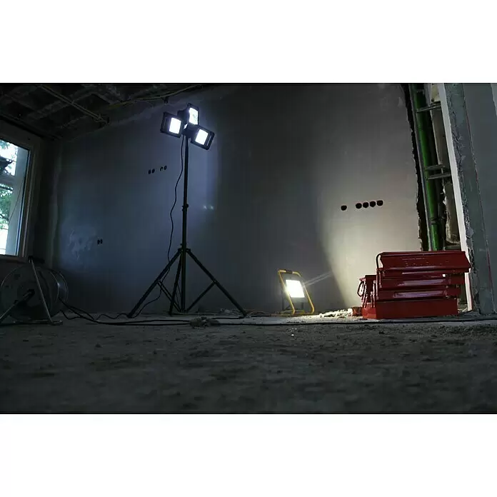 Profi Depot Proyector LED 2 cabezales - 50 W, 8.000 lm, 6.000 K - 7.000 K, L x An x Al: 1,14 x 1,32 x 2,25 m, IP54 - Dos focos de trabajo iluminan una habitación sin terminar. Uno grande con trípode y uno pequeño amarillo en el suelo, junto a una caja de herramientas roja.